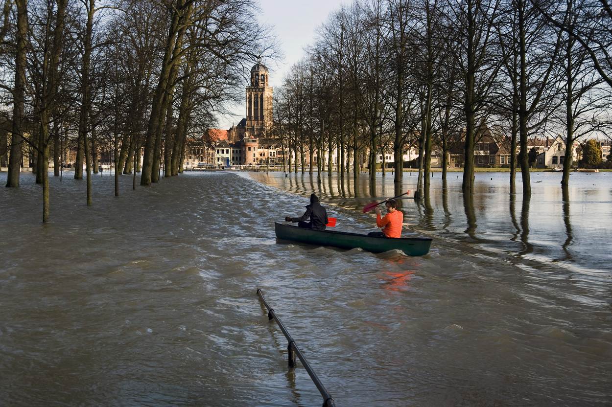 Twee mensen kanoën op een overstroomde weg. In de achtergrond is een dorp.